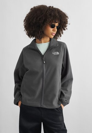 Person med krøllete hår som har på seg runde svarte solbriller, en grå fleecejakke med glidelås og svarte bukser, stående med hendene i lommene.