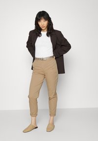 Brun blazer over en hvit skjorte, beige smale bukser og flettede brune slip-on sko. Outfiten har et sølvbelte og oppbrettede ermer.