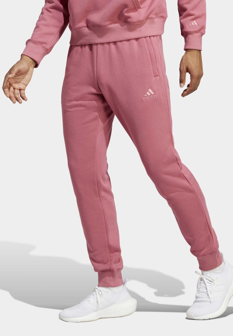 Rosa Jogginghose aus weichem Material, mit schmalem Schnitt, elastischem Bund und Seitentaschen. Das Adidas-Logo ist in Weiß aufgedruckt.