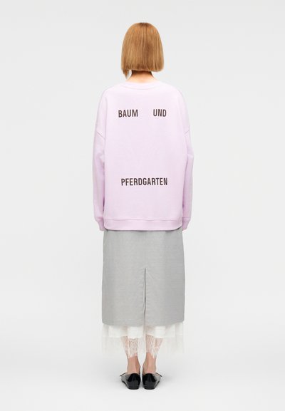 Ροζ ανοιχτόχρωμο φαρδύ φούτερ με μαύρο κείμενο «BAUM UND PFERDGARTEN» στο πίσω μέρος, συνδυασμένο με γκρι φούστα και μαύρα παπούτσια τύπου slip-on.