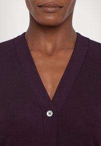 Gros plan sur le cou et le haut de la poitrine d'une personne portant un haut à col en V violet foncé fermé par un bouton blanc.