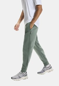 Lichtgroene joggers met zijzakken, elastische boorden en een subtiele donkere accent op de tailleband, gecombineerd met grijze sportschoenen.