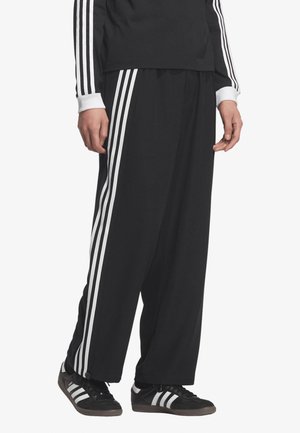 Persona che indossa pantaloni sportivi neri a gamba larga e top abbinato con dettaglio a tre strisce bianche, abbinati a scarpe da ginnastica nere Adidas.