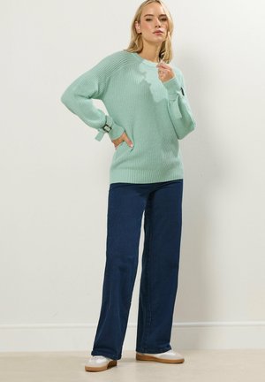 Blondynka w miętowo-zielonym swetrze z dzianiny z ozdobnymi klamrami, ciemnoniebieskich jeansach o szerokich nogawkach i białych trampkach na tle gładkiej ściany.