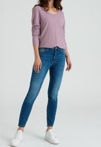 Světlofialový pletený svetr s výstřihem do V a dlouhými rukávy, doplněný modrými úzkými džínami a bílými teniskami. Textura působí měkce a neformálně.