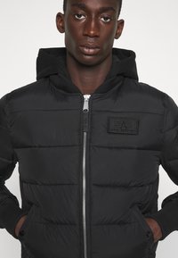 Schwarze Puffjacke mit Reißverschluss vorne, Kapuze und gerippten Bündchen. Besitzt eine gesteppten Struktur und ein auffälliges schwarzes Markenlogo auf der Brust.