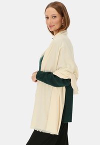 Kremowy szal o teksturowym wykończeniu noszony na ciemnozielonym swetrze. Szal ma frędzlowe krawędzie i lekko zwiewny krój.