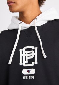 Felpa nera con cappuccio e cordino bianco, caratterizzata da un grande logo bianco e il testo "ATHL. DEPT.". Realizzata in un tessuto testurizzato con un cappuccio bicolore.