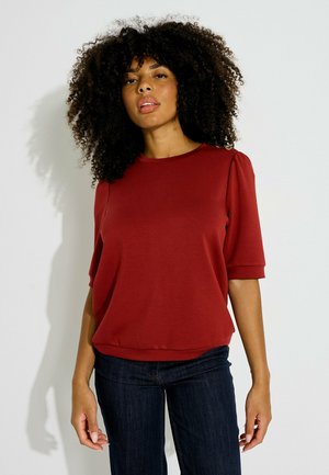 Femme aux cheveux bouclés portant un haut rouge à manches courtes et un jean foncé, debout devant un fond clair uni.