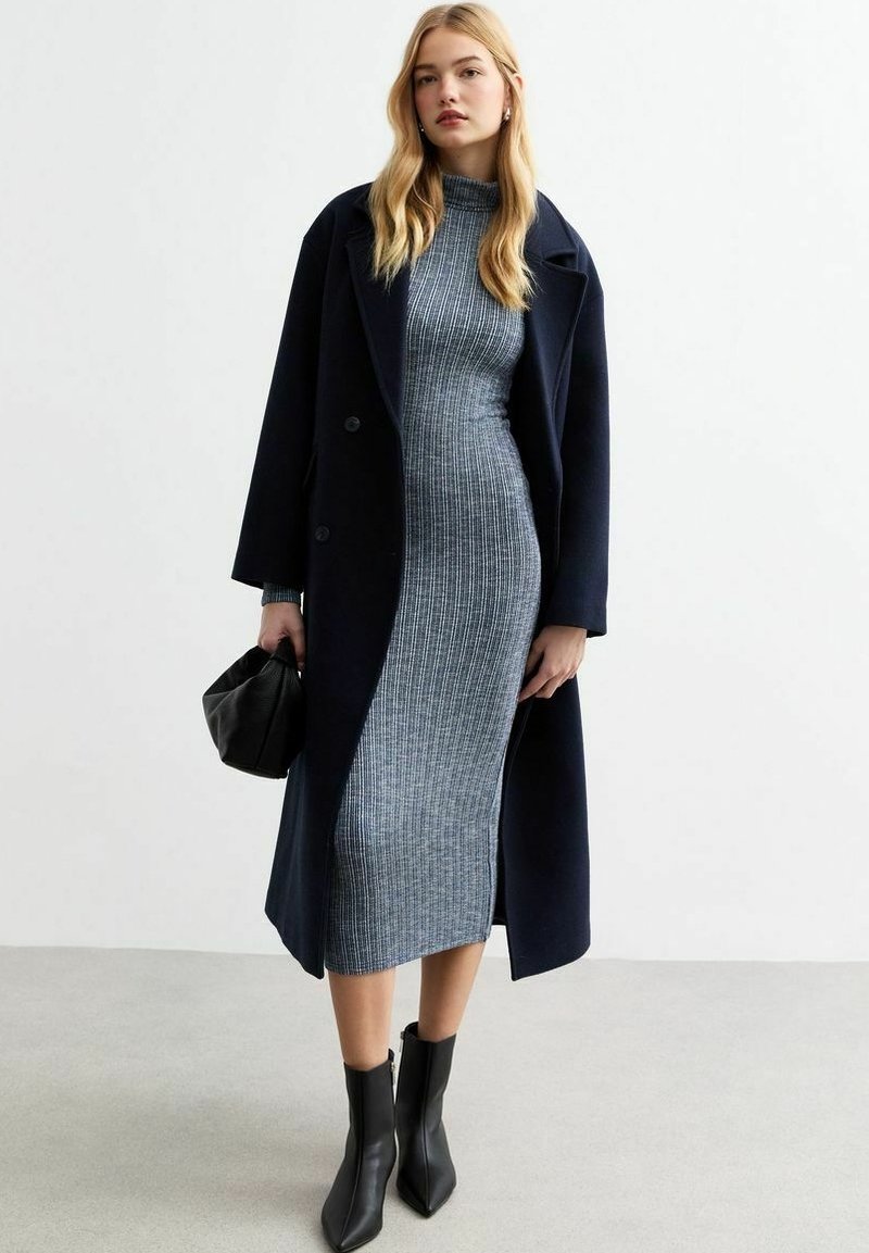 Cappotto navy con un design strutturato su un abito grigio fitted e a costine. Gli accessori includono una borsa nera e stivaletti neri con punta.