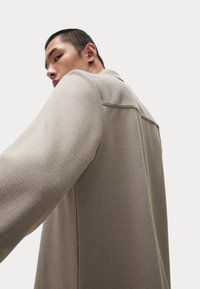 Veste en maille beige avec une surface texturée, des manches longues et une coupe droite. Le col est cranté et des coutures sont visibles le long du dos.