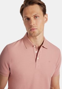 Jasnoróżowa koszulka polo z krótkim rękawem, dopasowana, z trzykrotnym zapięciem guzikowym oraz subtelnym logo na klatce piersiowej. Wykonana z teksturowanej bawełny.
