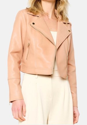 Donna che indossa una giacca corta in pelle beige con tasche con zip sopra un top chiaro e pantaloni crema a vita alta su sfondo bianco.