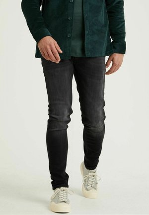 Mand iført mørkegrøn kordskjorte, sorte jeans og hvide sneakers står foran en ensfarvet baggrund.