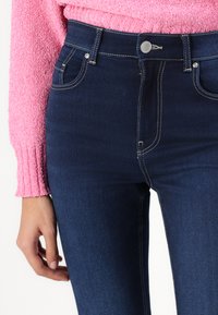 Jeans din denim albastru închis au un croi ajustat, talie înaltă și cusături contrastante. Se observă un sistem de închidere cu un buton argintiu.