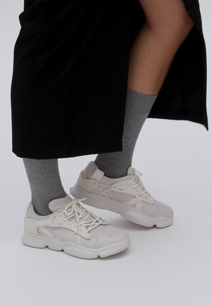 White sneakers with layered design, textured panels, and a chunky sole. Paired with ribbed grey socks, in a neutral setting.