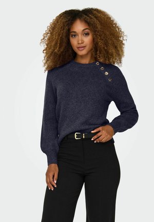 Femme aux cheveux bouclés portant un pull bleu foncé avec des boutons aux épaules et un pantalon noir, posant avec une main sur la hanche.
