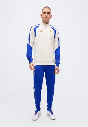 Uomo in piedi che indossa una giacca sportiva bianca e blu della Juventus e pantaloni della tuta blu con scarpe da ginnastica bianche su uno sfondo semplice.