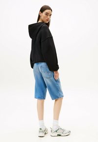 Sudadera negra de gran tamaño con capucha, combinada con pantalones cortos de mezclilla azul claro. El modelo lleva calcetines blancos y zapatillas multicolores.