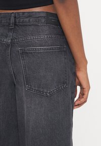 Jeans de denim cinza-escuro com um bolso traseiro, detalhes cosidos e uma etiqueta de marca na cintura. Textura suave e corte reto.
