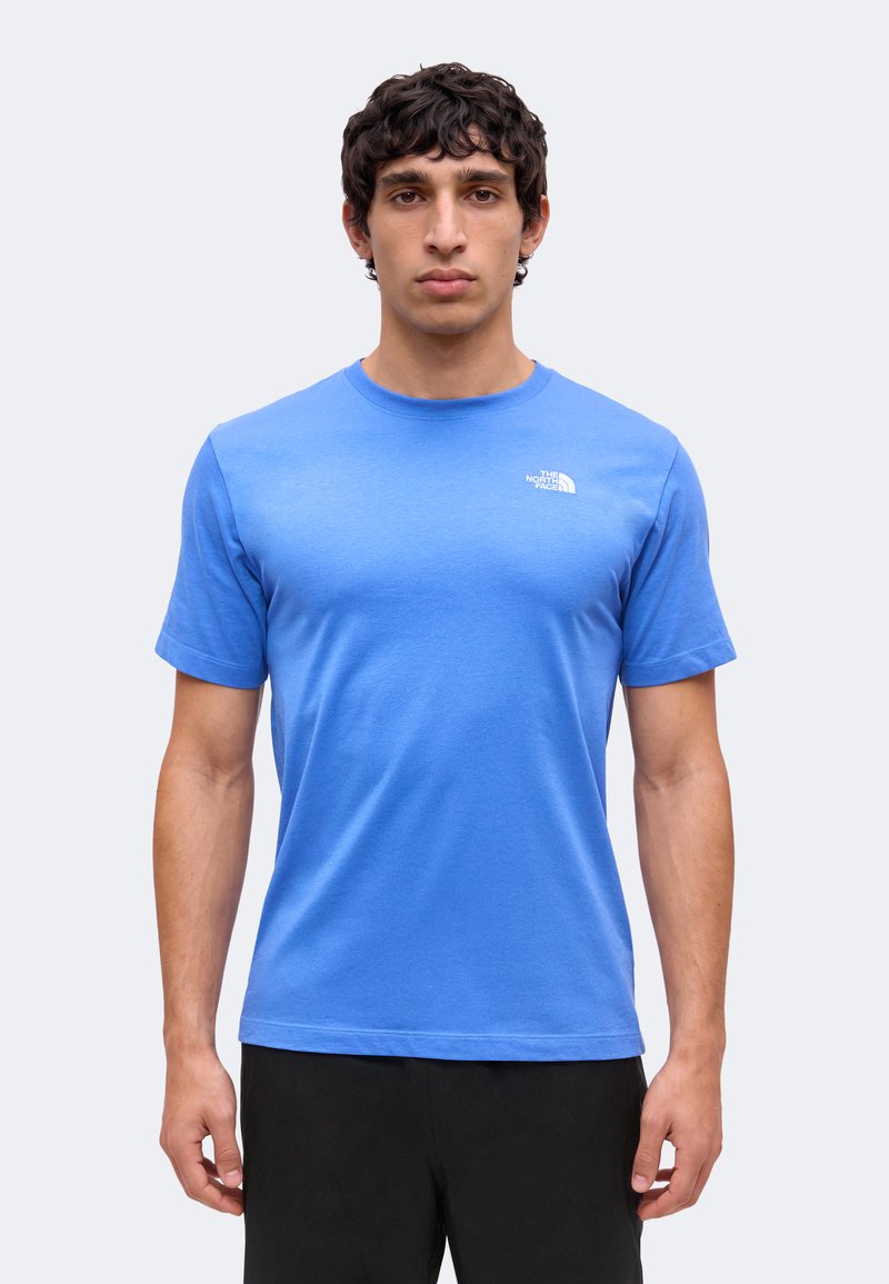 Hombre con cabello corto y rizado, que lleva una camiseta azul de The North Face y pantalones negros, de pie frente a un fondo claro y liso.