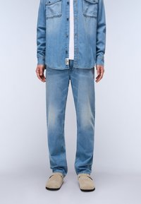 Camisa de denim azul claro con dos bolsillos en el pecho, combinada con pantalones vaqueros azul claro desgastados y zapatos slip-on beige. Fondo liso.
