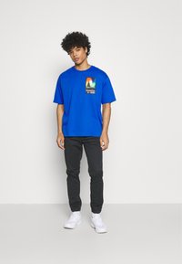 T-shirt en coton bleu avec un graphique carré sur la poitrine gauche présentant des couleurs arc-en-ciel et du texte ; associé à un pantalon noir ajusté et des baskets blanches.