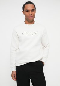 Hombre vistiendo un sudadera blanca de Guess y pantalones negros, de pie con una mano en el bolsillo contra un fondo blanco liso.