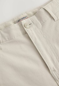 Pantalon en coton beige avec fermeture à bouton, présentant des détails cousus et une étiquette de marque à l'intérieur de la ceinture.