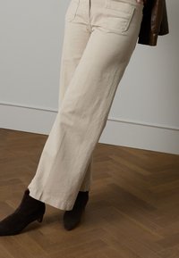 Beige Hose mit weitem Bein aus weichem Stoff, zwei Gesäßtaschen, kombiniert mit dunkelbraunen Stiefeletten auf einem Fischgrät-Holzboden.