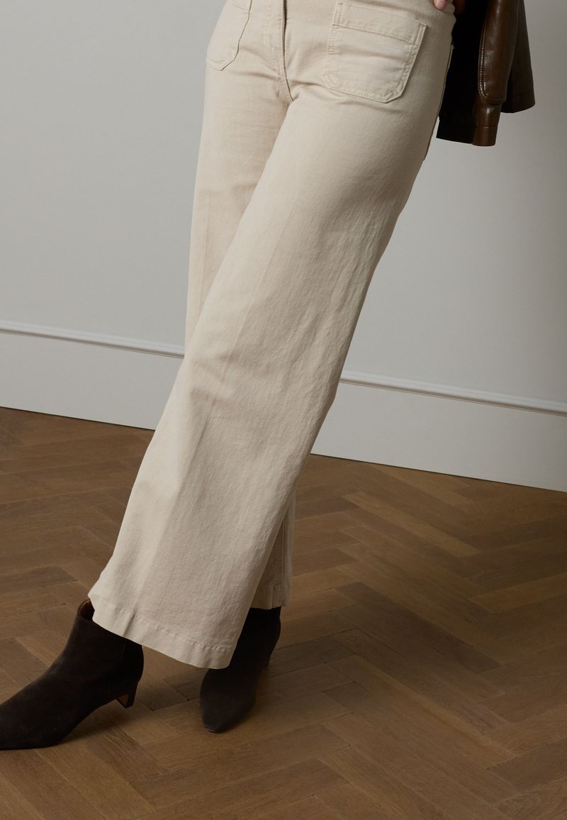 Beige Hose mit weitem Bein aus weichem Stoff, zwei Gesäßtaschen, kombiniert mit dunkelbraunen Stiefeletten auf einem Fischgrät-Holzboden.