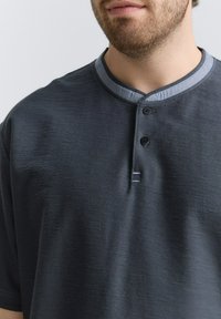 Donkergrijze T-shirt met korte mouwen van een gestructureerde stof, met een blauwe kraag en drie zwarte knopen aan de voorkant voor sluiting.
