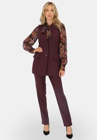 Fioletowa kamizelka z wyciętymi klapami, połączona z wzorzystą bluzką w intensywnych kolorach i długimi rękawami oraz pasującymi spodniami.