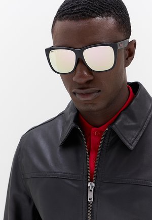 Uomo con occhiali da sole riflettenti, giacca di pelle nera con cerniera e camicia rossa con colletto su sfondo uni.