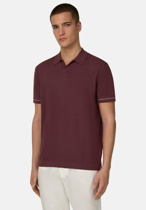 Jeune homme portant un polo bordeaux à manches courtes avec un liseré blanc sur les manches et un pantalon blanc, debout devant un fond uni.