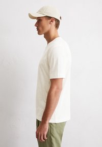 Hellbeige Baumwoll-T-Shirt mit kurzen Ärmeln und Rundhalsausschnitt, kombiniert mit hellgrünen Hosen. Einfaches Design, weiche Textur und minimale Details.