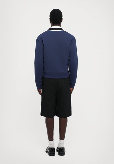 Navy blue sweatshirt with textured fabric, white collar detailing, and a cropped design, paired with black shorts and black shoes.