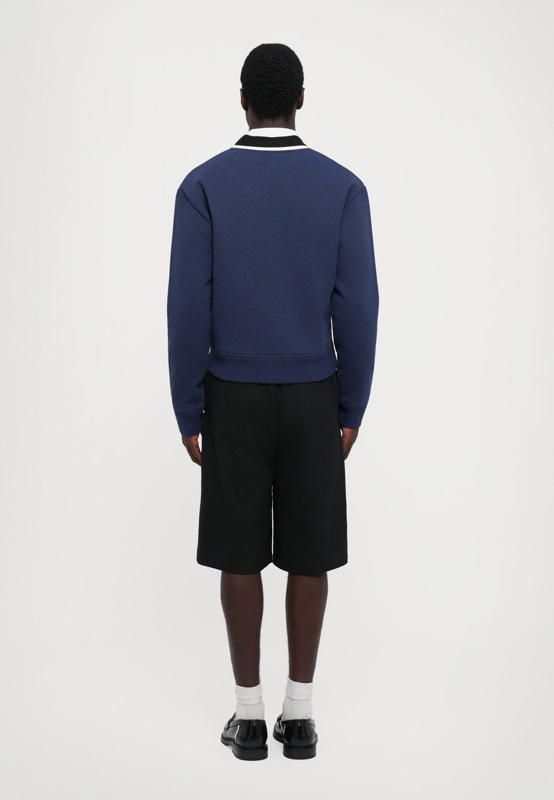Sudadera azul marino con tejido texturizado, detalles en el collar blancos y un diseño corto, combinada con pantalones cortos negros y zapatos negros.