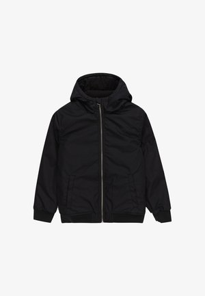 Jeune personne portant une veste à capuche noire, un jean bleu large et des baskets noires avec des semelles en gomme, les mains dans les poches, debout sur un fond blanc.