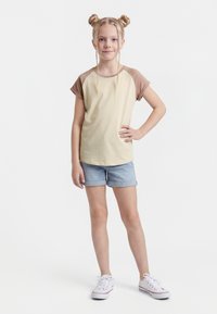 Ung jente med blondt hår i to boller og fletter, iført beige og brun t-skjorte, denimshorts og hvite joggesko, stående med hånden på hoften.