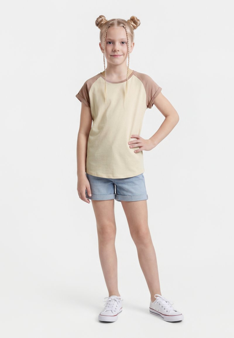 Ung jente med blondt hår i to boller og fletter, iført beige og brun t-skjorte, denimshorts og hvite joggesko, stående med hånden på hoften.