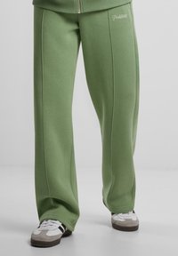 Groene, losse sweatpants van zachte stof, met rechte pijpen en een logo op de linkerheup. Gecombineerd met grijs-witte sneakers.