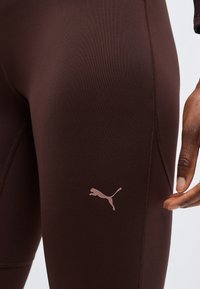 Κοντινό πλάνο σκούρων καφέ αθλητικών κολαν με ορατές ραφές και ελαφρώς καφέ λογότυπο Puma στον μηρό δίπλα σε ένα χέρι.