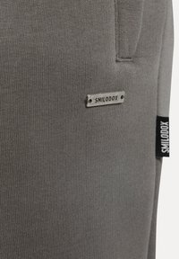 Tela gris acanalada con una prominente etiqueta metálica con el logo de Smilodox. Presenta un detalle de costura lateral con una etiqueta negra. Diseño ajustado.