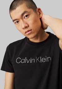 T-shirt en coton noir avec un logo "Calvin Klein" brodé en gris sur le devant, manches courtes et col rond.