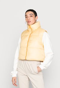 Colete acolchoado amarelo cropped com gola alta e fecho dianteiro. Usado sobre uma camisa branca de mangas longas, combinado com calças azul claro.