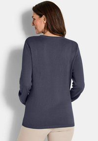 Frau mit braunen Haaren, die einen langärmeligen, dunkelgrauen Pullover und beige Hosen trägt, von hinten vor einer weißen Wand gezeigt.