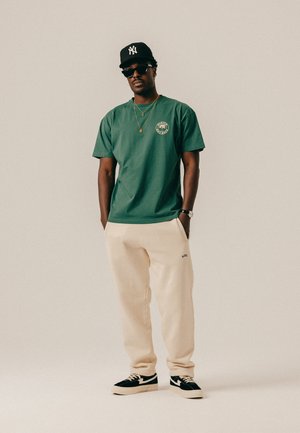 Mand iført grøn t-shirt, beige joggingbukser, sort New York Yankees kasket, solbriller, guldhalskæder og sorte sneakers, stående med hænderne i lommerne.