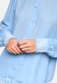 Light blue blouse featuring white polka dots, button closure, and gathered sleeves. Cuffs adorned with delicate white lace trim.