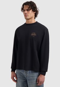 Mann mit lockigem Haar, der ein schwarzes Langarmshirt mit einem kleinen beige Logo auf der Brust und hellblauen Jeans trägt, vor einem weißen Hintergrund steht.